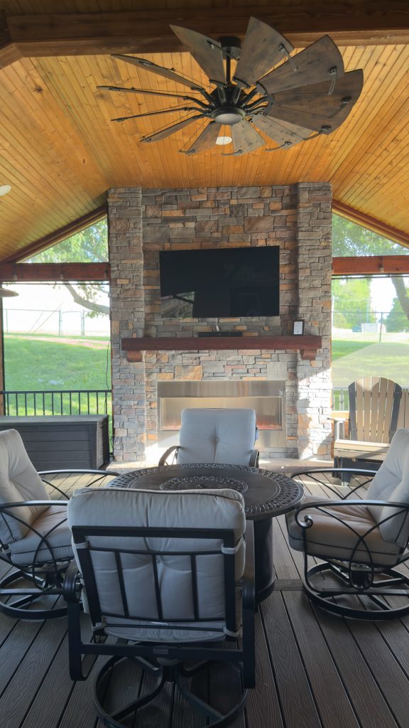 Central seating area on a Kansas City screened porch featuring a large stone fireplace, mounted TV, and a round fire pit table.